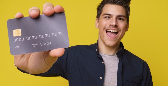 Impressed handsome young person holds in hand debit credit card happy open mouth isolated on yellow color background. Close up of big bank card in hand of 20s young man hipster in blue shirt