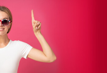 Cute blond beautiful woman half portrait wearing glasses dressed in white t-shirt raise pointing finger up, showing promo, give direction, isolated pink red background. Banner woman attention notes.