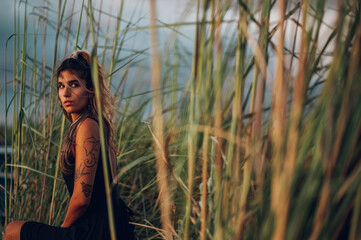 Portrait of a young fashionable tanned woman with tattoos sitting in nature surrounded by grass.