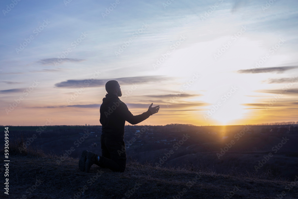 Wall mural A man with raised hands is praying. On the background of the sunset sky. Repentance for sins. Kneeling Prayer to God. Worship and praise.