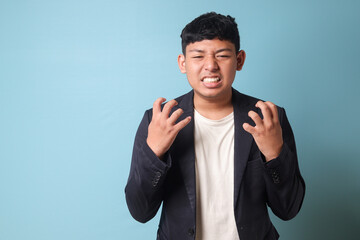 Portrait of young Asian business man in casual suit holding back anger. Isolated image on blue background