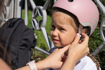 Mother putting on inline skates rollers protection helmet to daughter child in public park in...