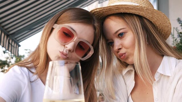 Two young beautiful smiling hipster female. Carefree women posing at veranda cafe in the street. Positive models drinking white wine. Enjoying their vacation. Eating croissant. Taking selfie