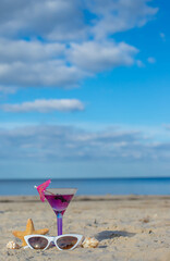 Glass of cocktail on sandy beach and sunglasses next to sea