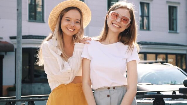 Two young beautiful smiling hipster female in trendy summer clothes. carefree women posing on the street background in hat. Positive pure models having fun at sunset, hugging 