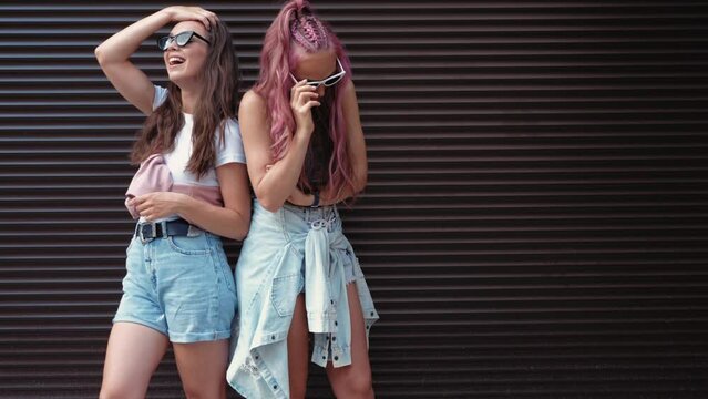 Two young beautiful smiling hipster female in trendy summer clothes.Sexy carefree women posing in the street near roller shutter wall with pink hair. Positive pure models in sunglasses
