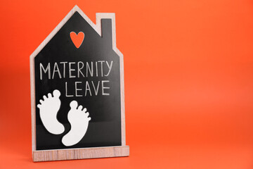 Wooden house figure with words Maternity Leave and paper cutout of baby feet on orange background. Space for text