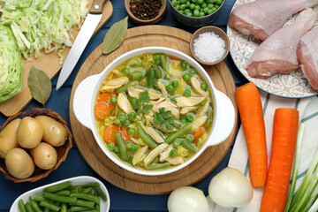Saucepan of delicious vegetable soup with chicken and different ingredients on blue wooden table, flat lay