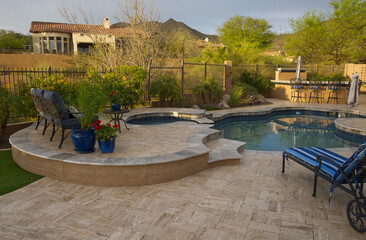Fototapeta premium A desert landscaped backyard in Arizona featuring a travertine tiled pool deck with spa and outdoor kitchen.