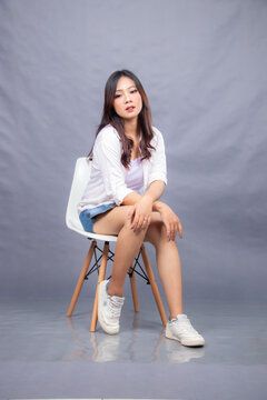 Elegant Woman Posing Sitting On A Chair In Front Of The Camera While Her Ladder Leans Forward Indoors. Fashion Promotion Women's Shirt Photo Session In Gray Background Studio