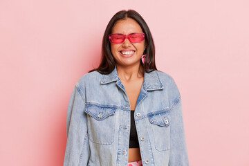 Young pretty girl in denim jacket and pink sunglasses who stands in the middle of pink wall in the...