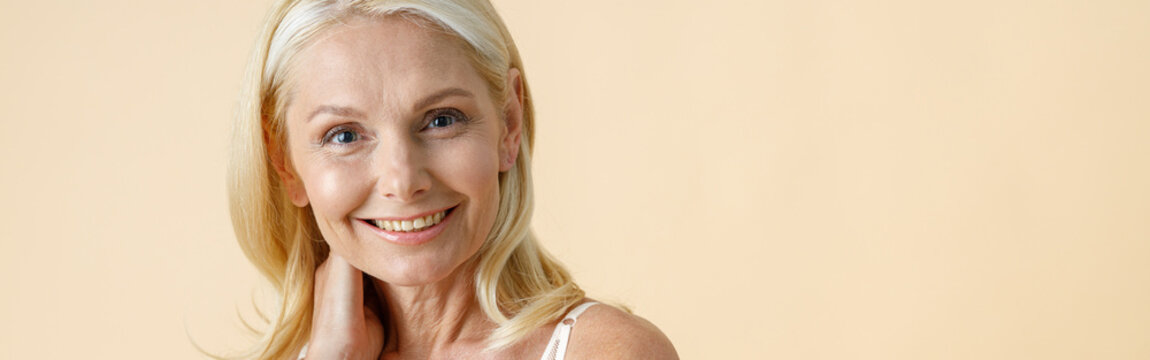 Portrait of charming mature woman with blonde hair in white underwear smiling at camera, posing isolated over beige background