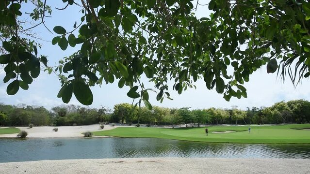 Video at a golf course in tropical Mexico with blue skies, lush foliage, and a man-made lake. Two players on the green execute the compo for birdie
