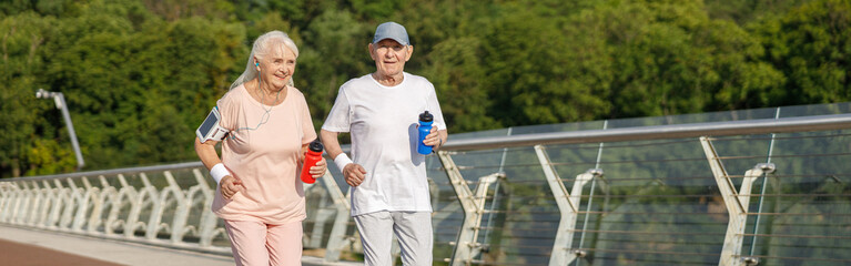 Positive seniot couple with bottles of water runs training together on footbridge