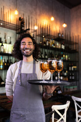 Vertical portrait of happy waiter standing in bar holding tray of beer looking at camera.