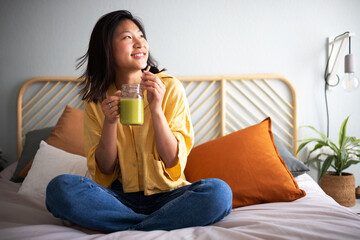 Pensive, happy young asian woman sitting on bed drinking green juice smoothie.