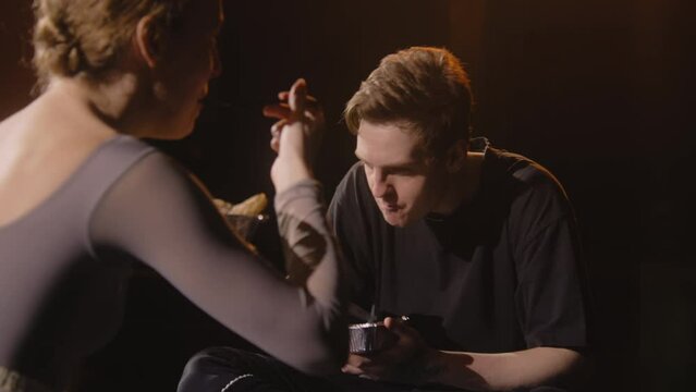 Pair of tired ballet dancers sit on theatre stage and discuss performance. Ballerina and dance partner eat together after choreography rehearsal. Empty theatrical hall. Classical ballet art concept.