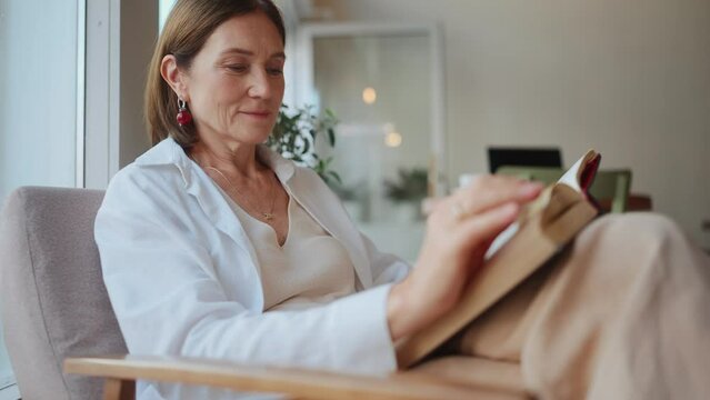 Pensive elderly woman in white shirt reading book in cafe