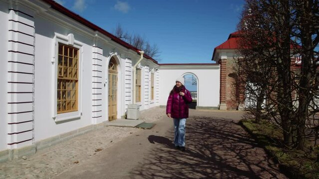 Tourist woman walking in yard and looking at white baroque style one-storey building early spring in touristic place. Female wearing jacket hat jeans. Tourism, travel, vacations, sightseeing concept. 