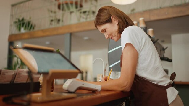 Cute aged woman barista signs paper documents behind counter in cafe