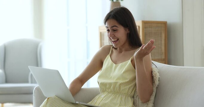 Focused teen student girl using online learning app on laptop, resting on home couch, getting good surprising news, shouting yes, making winner gesture, celebrating success, enjoying communication