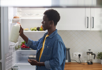 Side view portrait of young black man taking milk from refrigerator while cooking dinner at home