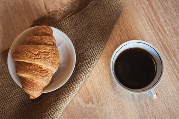A cup of hot fragrant black coffee with a croissant in a saucer on a cloth, on a light brown wooden table. An image as a background for your design and creative illustrations.