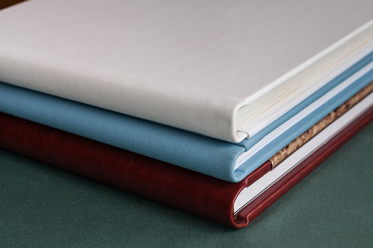 Three stylish photobooks with leather covers, white, burgundy and blue, of different thicknesses, lie on a dark green surface in a room.