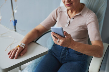 Minimal closeup of mature woman using smartphone during IV drip treatment, copy space