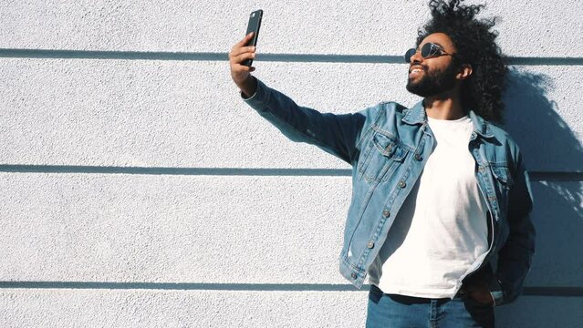 Handsome smiling hipster  model. Sexy unshaven Arabian man dressed in summer jeans jacket clothes. Fashion male with long curly hairstyle posing in the street at sunset. Taking selfie photos