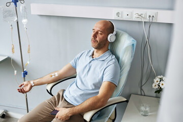 High angle portrait of adult man wearing headphones and relaxing during IV drip treatment in clinic