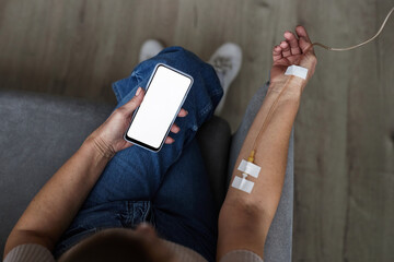 Top view closeup of unrecognizable man using smartphone with white screen mockup during IV drip treatment, copy space