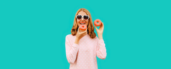 Portrait of happy cheerful laughing young woman with sweet donut having fun wearing heart shaped sunglasses on blue background