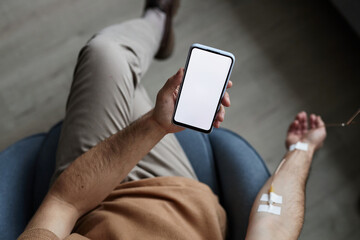 Top view of man using smartphone with white screen mockup during IV drip treatment, copy space