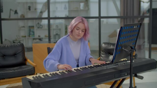 Talented beautiful pink haired woman pianist performing and playing electronic piano keyboard,, leafing pages with musical notes on sheet music stand while practicing in domestic room.