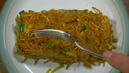 Closeup overhead shot of a man’s hands tipping a takeout carton of hot Chinese chicken and vegetable stir fried noodles onto a plate, then spreading them with a fork.