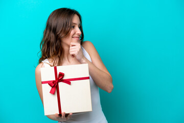 Young caucasian woman holding a gift isolated on blue background thinking an idea and looking side