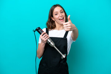 Young caucasian cooker woman using hand blender isolated on blue background with thumbs up because something good has happened