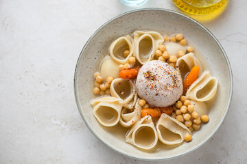 Plate of escudella with pasta, chickpeas and pilota, high angle view on a light-beige stone background, horizontal shot with space