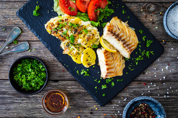Fried cod loin with baked potatoes and fresh vegetables on black plate on wooden table
