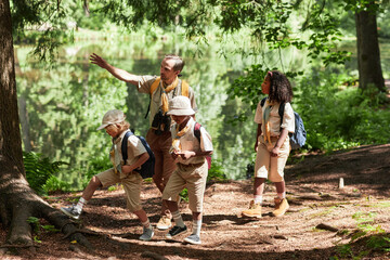 Side view at diverse group of scouts hiking in forest with adult leader pointing away in sunlight