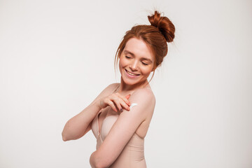 Happy beautiful young redhead fresh woman with cute smile in beige bra applying cream on her shoulder in studio on white background. Beauty, health and skin care
