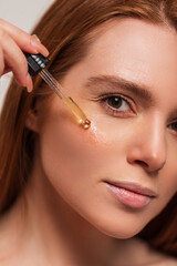 Beautiful young redhead woman with clean skin holds a pipette and drips oil on her face skin,...