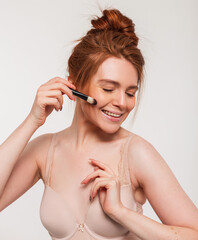 Fototapeta premium Happy beautiful young fresh redhead woman with a sweet smile doing makeup with a brush in the studio. Beauty and skin care