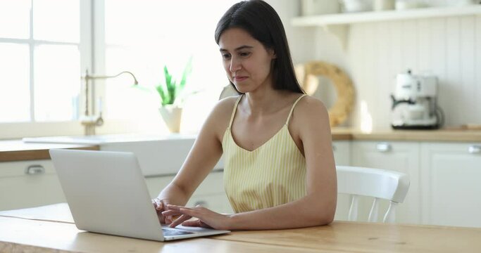 Happy positive Latin young adult girl using online app on laptop, working at computer, sitting at table in home kitchen, typing, smiling, laughing, looking away, thinking