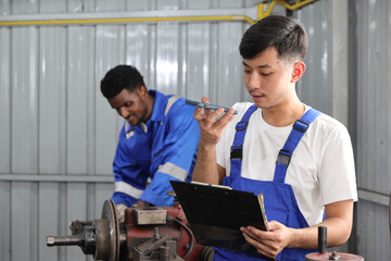 Multiethnic technicain mechanic using metal lathe machine operate polishing car disc brake with smart mobile phone and clipboard at garage. Maintenance automotive and inspecting vehicle part concept