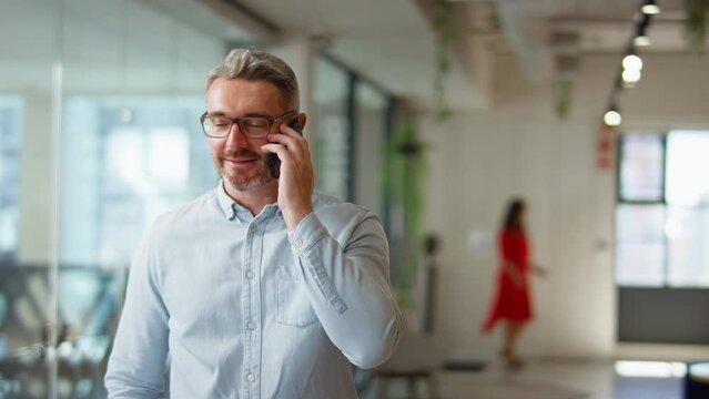 Camera tracks across mature businessman standing in modern open plan office talking on mobile phone -- shot in slow motion