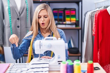 Naklejka na ściany i meble Blonde woman dressmaker designer using sew machine pointing down with fingers showing advertisement, surprised face and open mouth