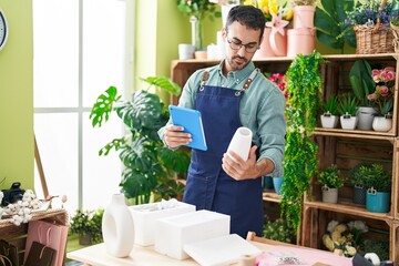 Young hispanic man florist using touchpad holding plant pot at flower shop