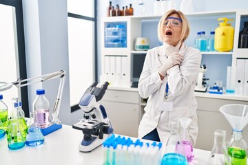 Middle age blonde woman working at scientist laboratory shouting suffocate because painful strangle. health problem. asphyxiate and suicide concept.